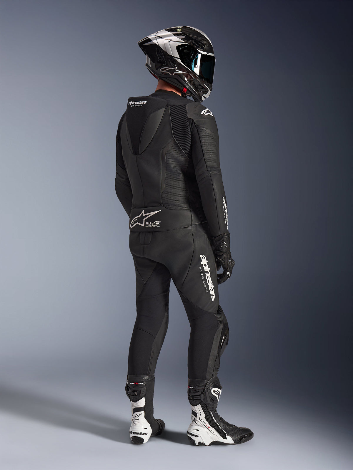 Alpinestars GP Force V2 2-Piece Leather Suit, Black, rear view of motorcycle rider wearing full racing gear, featuring aerodynamic speed hump, white branding, and perforated leather, paired with a carbon fiber helmet and black and white racing boots