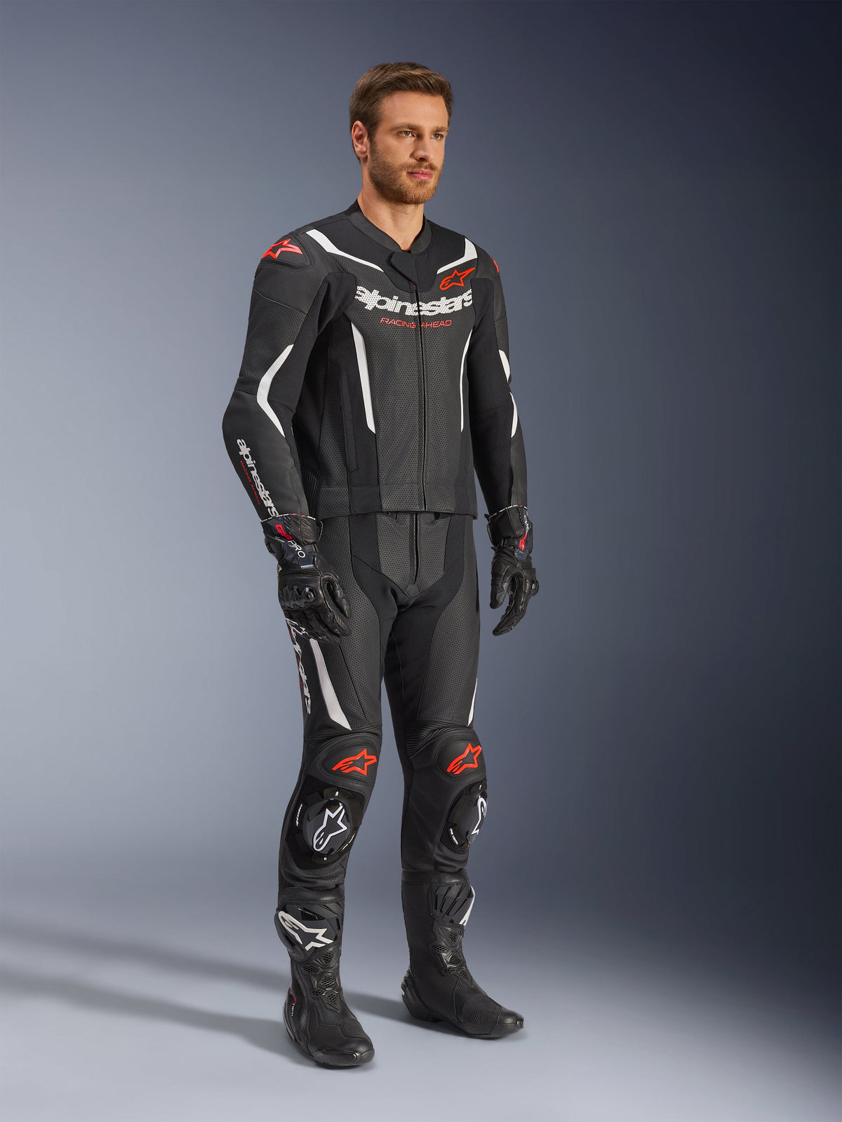 Fato de Pele Alpinestars GP Force V2 de 2 Peças, preto, branco e vermelho fluo, vestido por um modelo com botas e luvas de corrida pretas, com painéis de pele perfurada, detalhes aerodinâmicos brancos e logótipos vermelho fluo nos ombros e joelhos, equipamento profissional de motociclismo
