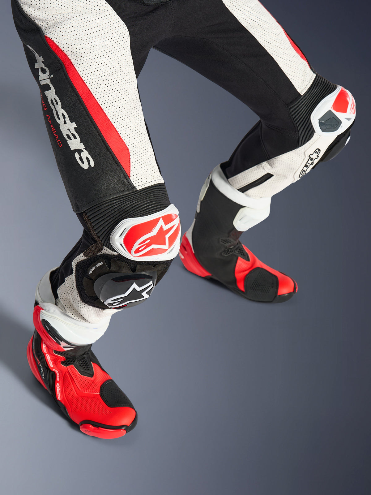 Alpinestars Stella GP-R7 2-Piece Leather Suit para Mulher, zona inferior das pernas e botas de corrida, em Preto Branco e Vermelho Fluo, com pele perfurada, logótipo Alpinestars proeminente na coxa, painéis de extensão em sanfona e deslizadores de joelho vermelhos e brancos com botas técnicas em vermelho e preto a condizer.