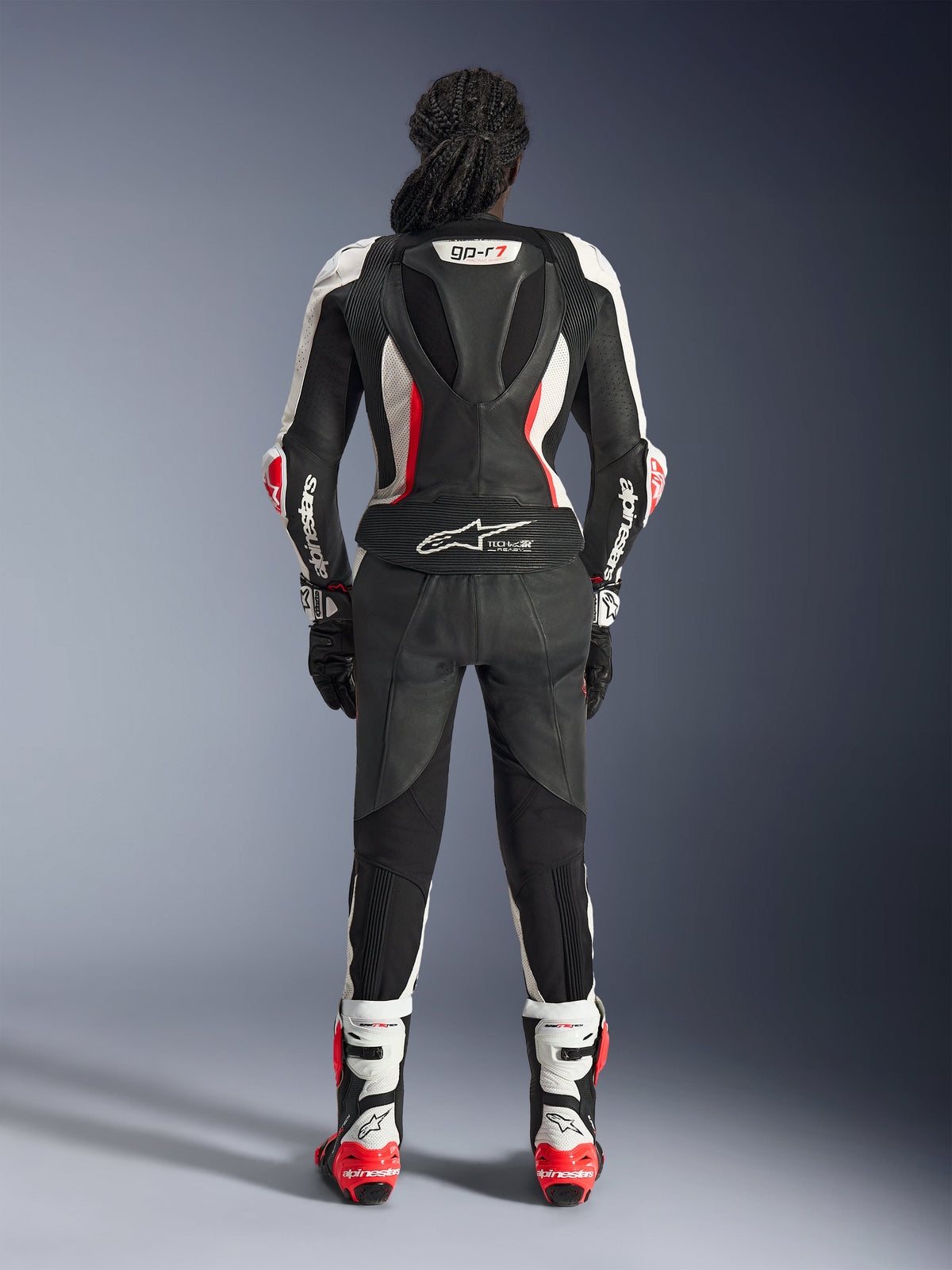 Vista traseira do fato de cabedal de 2 peças Alpinestars Stella GP-R7, preto, branco e vermelho fluo, com corcova aerodinâmica, marca GP-R7 e logótipo Tech-Air Ready, usado com botas e luvas de corrida a condizer, equipamento profissional de motociclismo para mulher