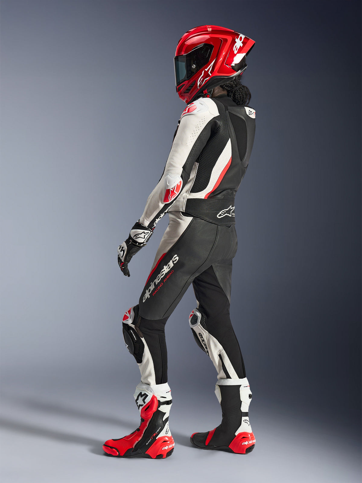 Motociclista feminina com fato de cabedal de 2 peças Alpinestars Stella GP-R7, preto, branco e vermelho fluo, vista de três quartos traseira a mostrar a corcova aerodinâmica, capacete integral vermelho a condizer, luvas de corrida pretas e brancas e botas de corrida técnicas vermelhas e brancas, equipamento de corrida profissional