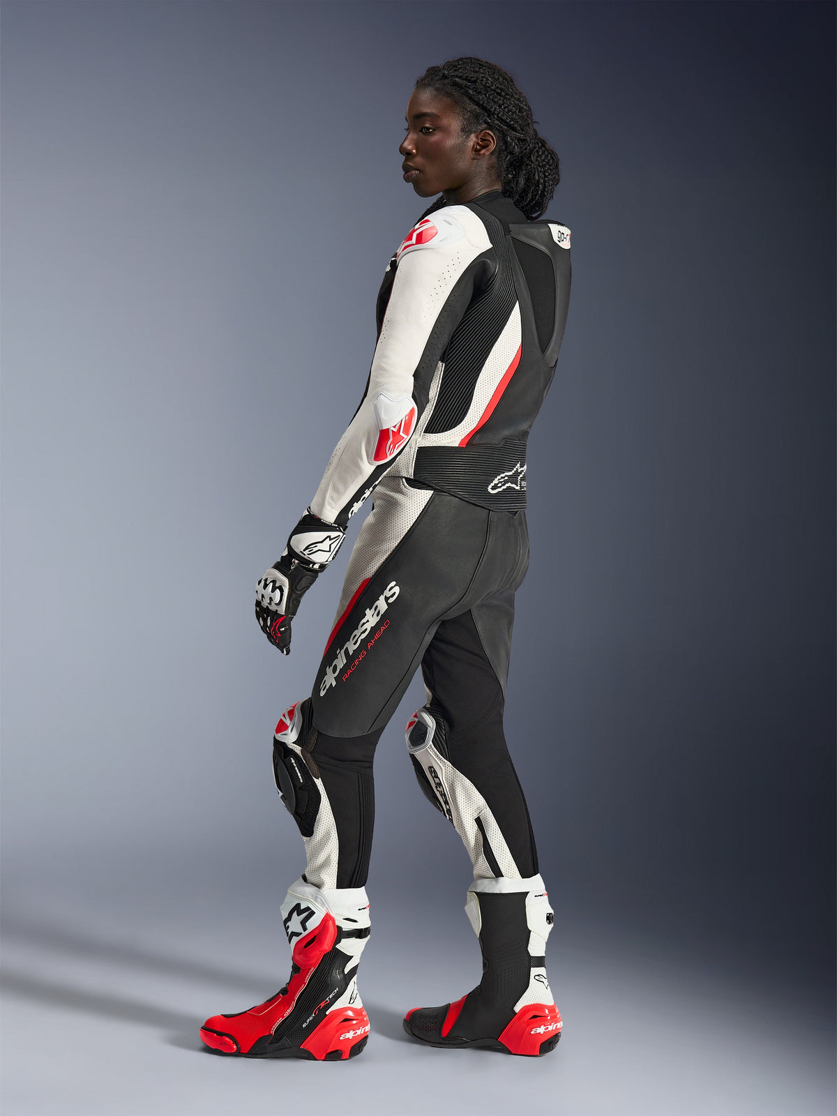 Mulher a usar o fato de cabedal de duas peças Alpinestars Stella GP-R7, em preto, branco e vermelho fluo, perfil lateral com corcova aerodinâmica e deslizadores de joelho, combinado com luvas e botas de corrida Alpinestars a condizer, equipamento profissional de pista para motociclismo