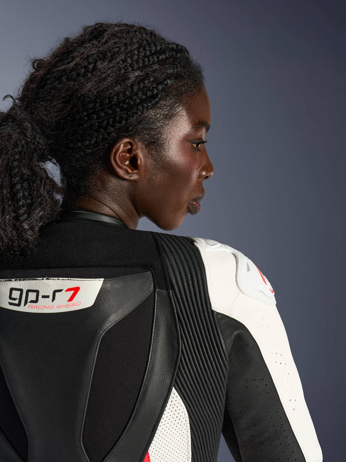 Perfil em grande plano de uma mulher a usar o fato de cabedal de 2 peças Alpinestars Stella GP-R7, mostrando a zona superior das costas e dos ombros em cabedal preto e branco, com o logótipo 'GP-R7', painéis elásticos em sanfona e proteção de ombro branca com um logótipo vermelho.