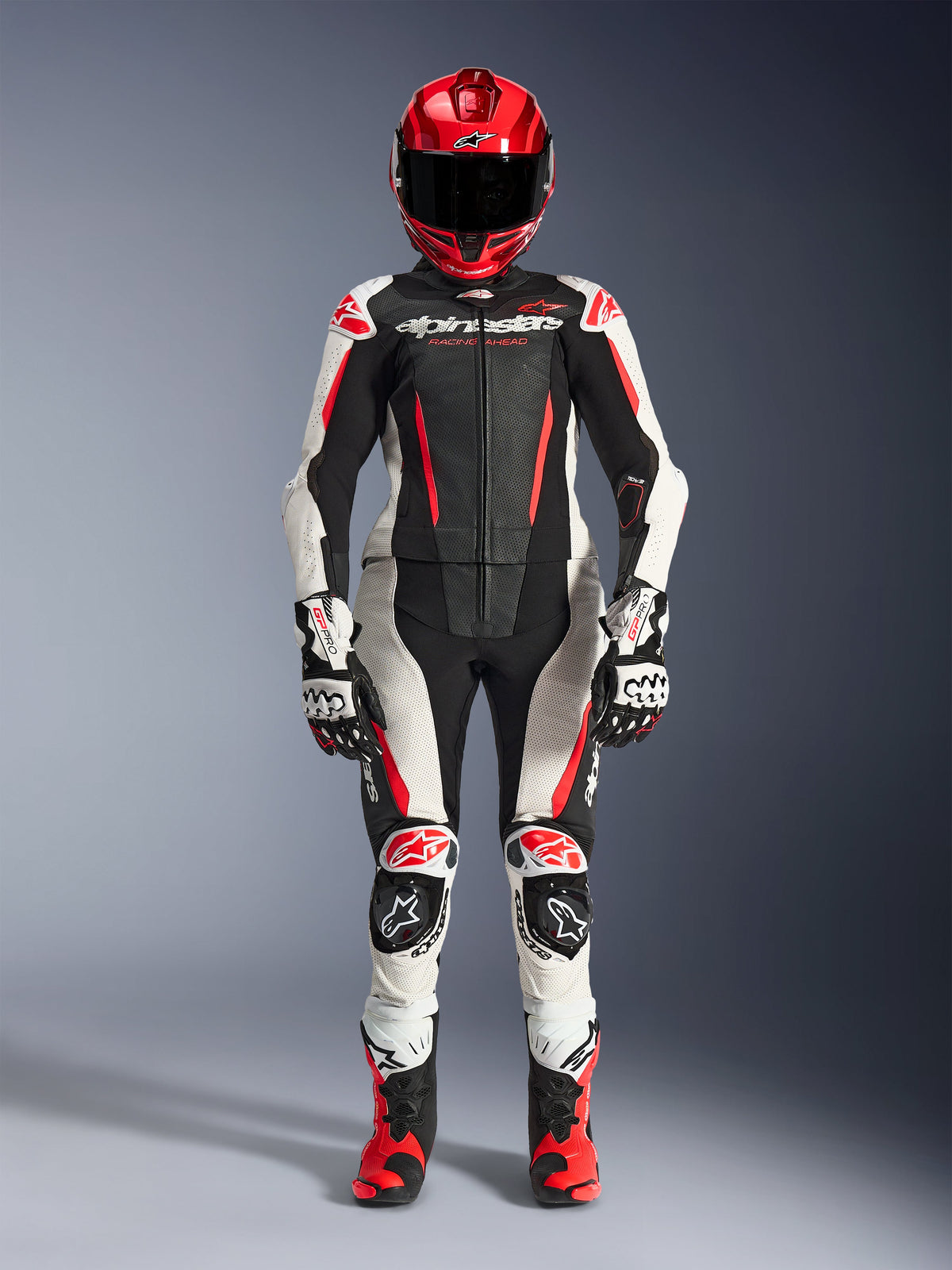 Fato de Cabedal de 2 Peças Alpinestars Stella GP-R7 para Mulher em preto, branco e vermelho fluo, vista de corpo inteiro de uma piloto a usar capacete, luvas e botas de corrida a combinar, com painéis perfurados e design aerodinâmico para condução em pista e estrada
