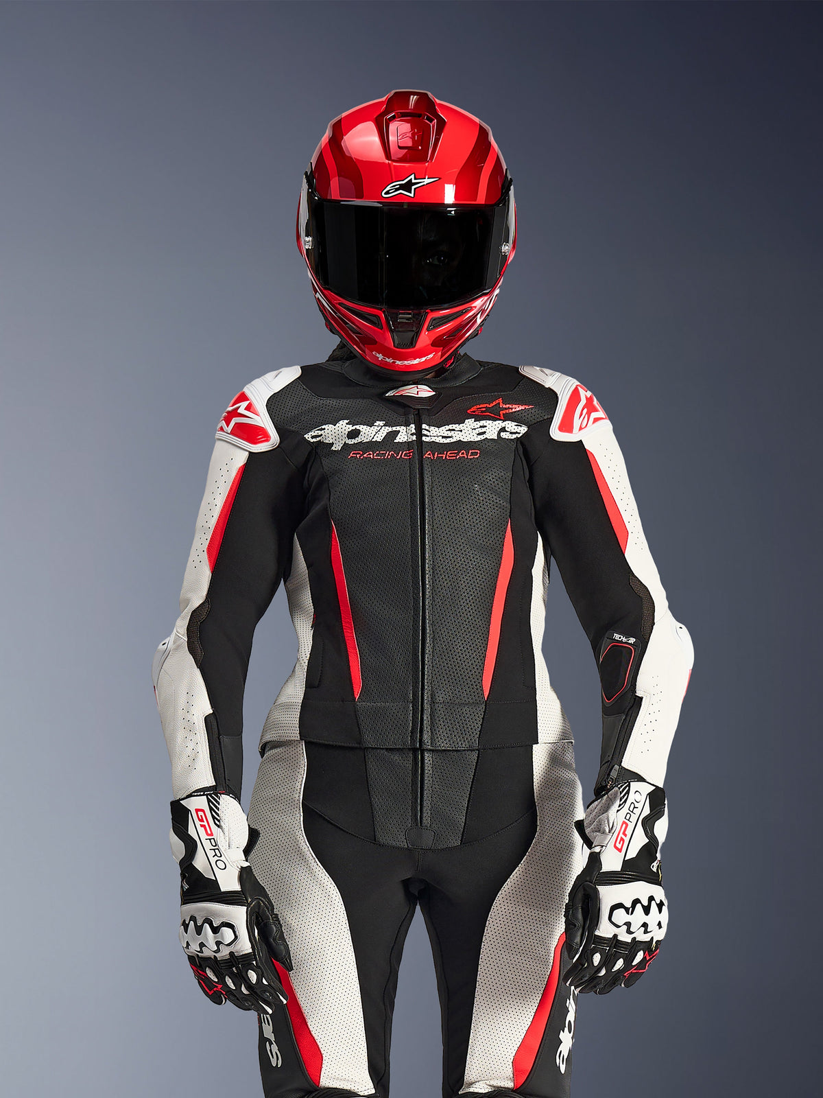 Piloto feminina com fato de cabedal de duas peças Alpinestars Stella GP-R7, nas cores preto, branco e vermelho fluo, com painéis perfurados e marca 'Racing Ahead', acompanhado de capacete vermelho e luvas GP Pro brancas e pretas, conjunto de equipamento de corrida profissional