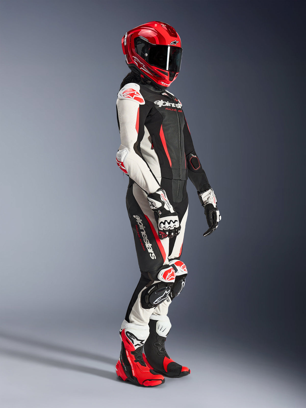 Motociclista a usar o fato de cabedal de 2 peças Alpinestars Stella GP-R7 em preto, branco e vermelho fluo, com capacete integral de corrida vermelho, luvas a condizer e botas de performance, equipamento profissional de motociclismo para mulher.