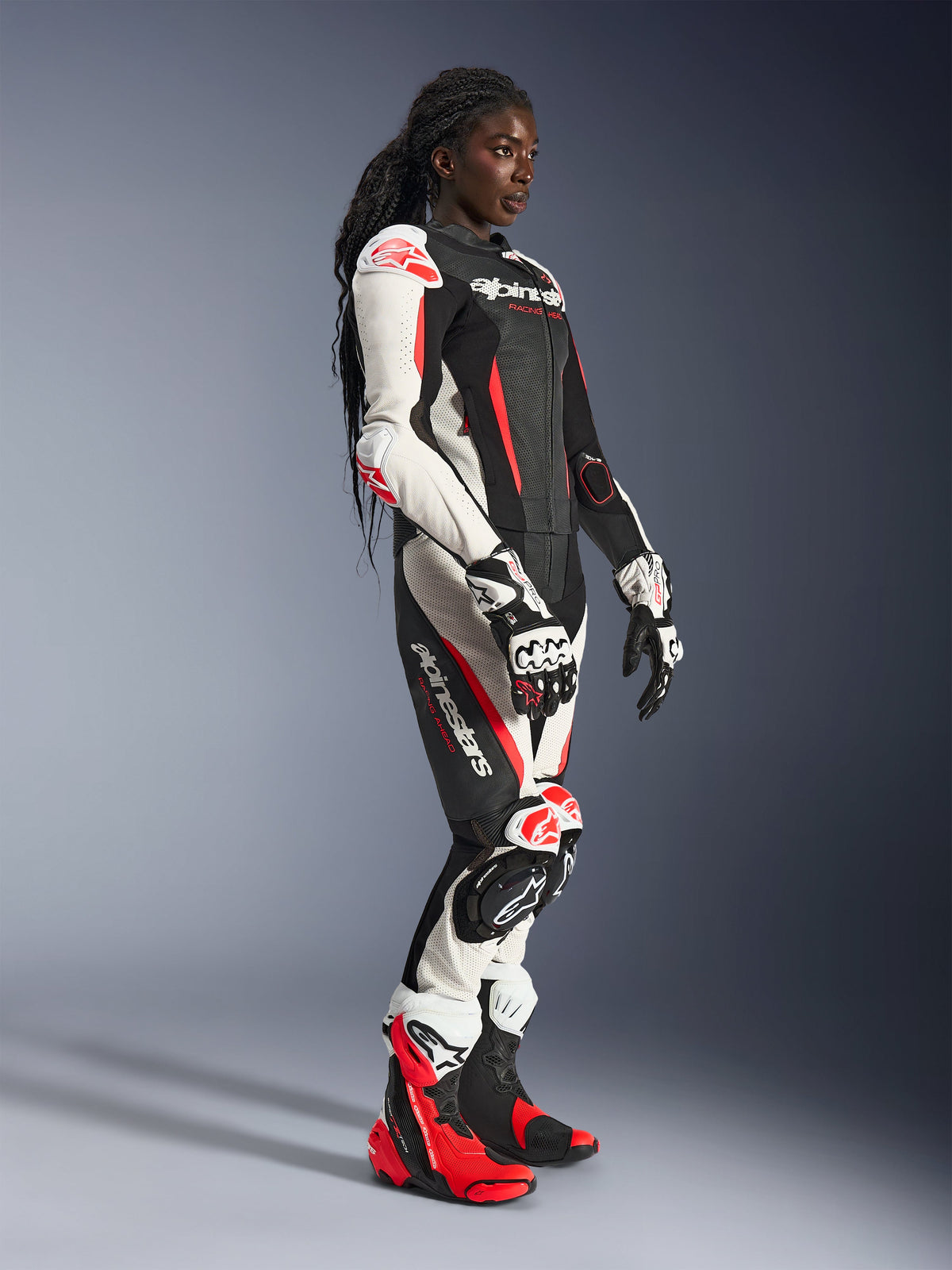 Motociclista a usar o fato de cabedal de 2 peças Alpinestars Stella GP-R7 em preto, branco e vermelho fluo, combinado com luvas e botas de corrida a condizer, com painéis de cabedal perfurado e proteções nos ombros, cotovelos e joelhos, em ambiente de estúdio.