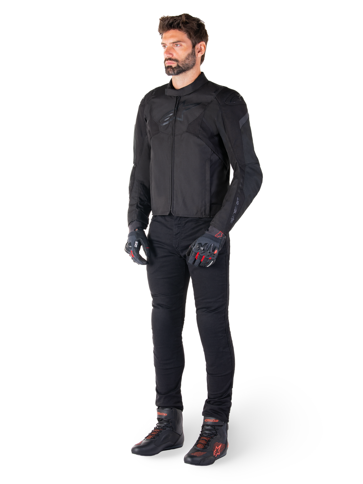 Modelo masculino a usar o Casaco Impermeável Alpinestars T-Jaws V4 em preto, combinado com calças de motociclismo pretas, luvas pretas com detalhes vermelhos e sapatos de motociclismo pretos, com um ajuste desportivo elegante e logótipos discretos, isolado num fundo branco.