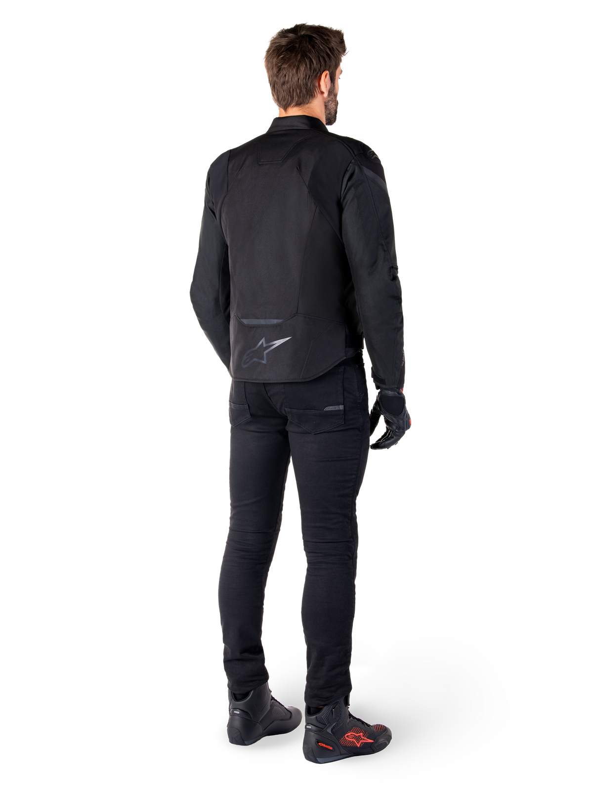 Casaco Impermeável Alpinestars T-Jaws V4, preto, vista traseira num modelo a usar calças e botas de motociclismo pretas, exibindo a construção têxtil desportiva e o logótipo Alpinestars na parte inferior das costas.