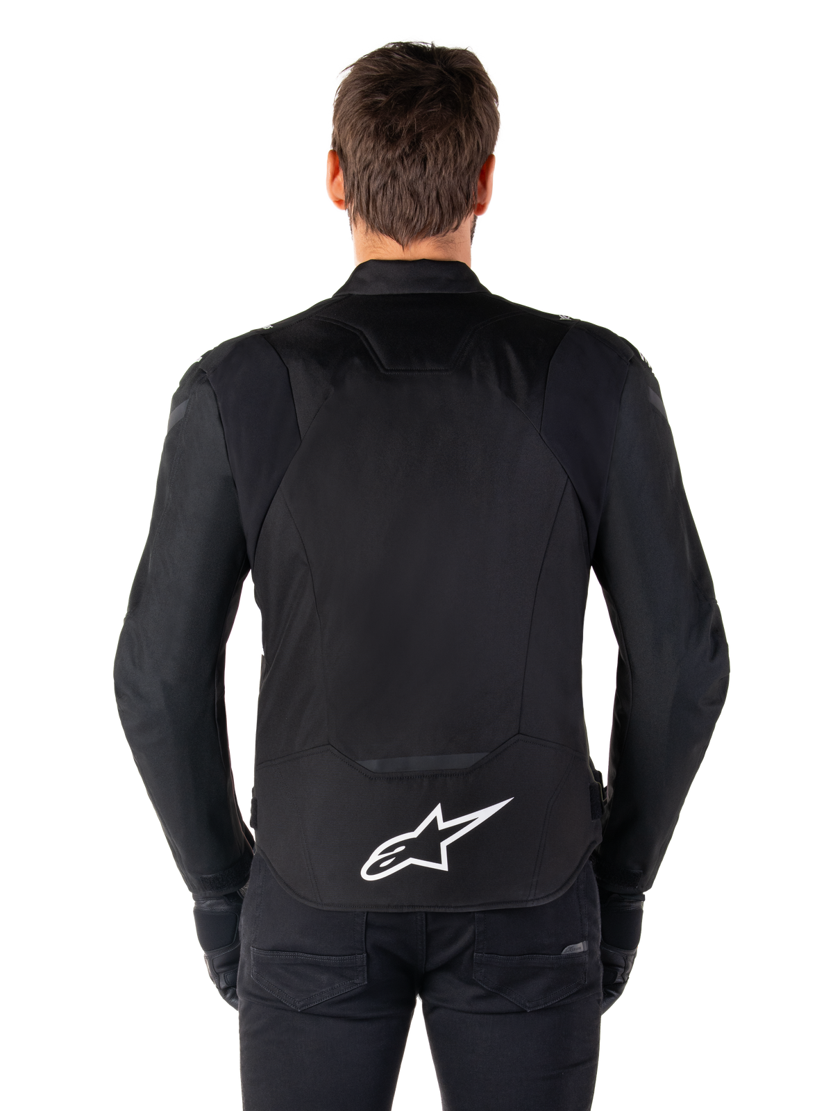 Casaco Impermeável Alpinestars T-Jaws V4, casaco de motociclismo preto visto de costas, com logótipo Alpinestars branco na parte inferior das costas, parte traseira prolongada para maior cobertura, usado por modelo com calças e luvas pretas, design têxtil desportivo elegante