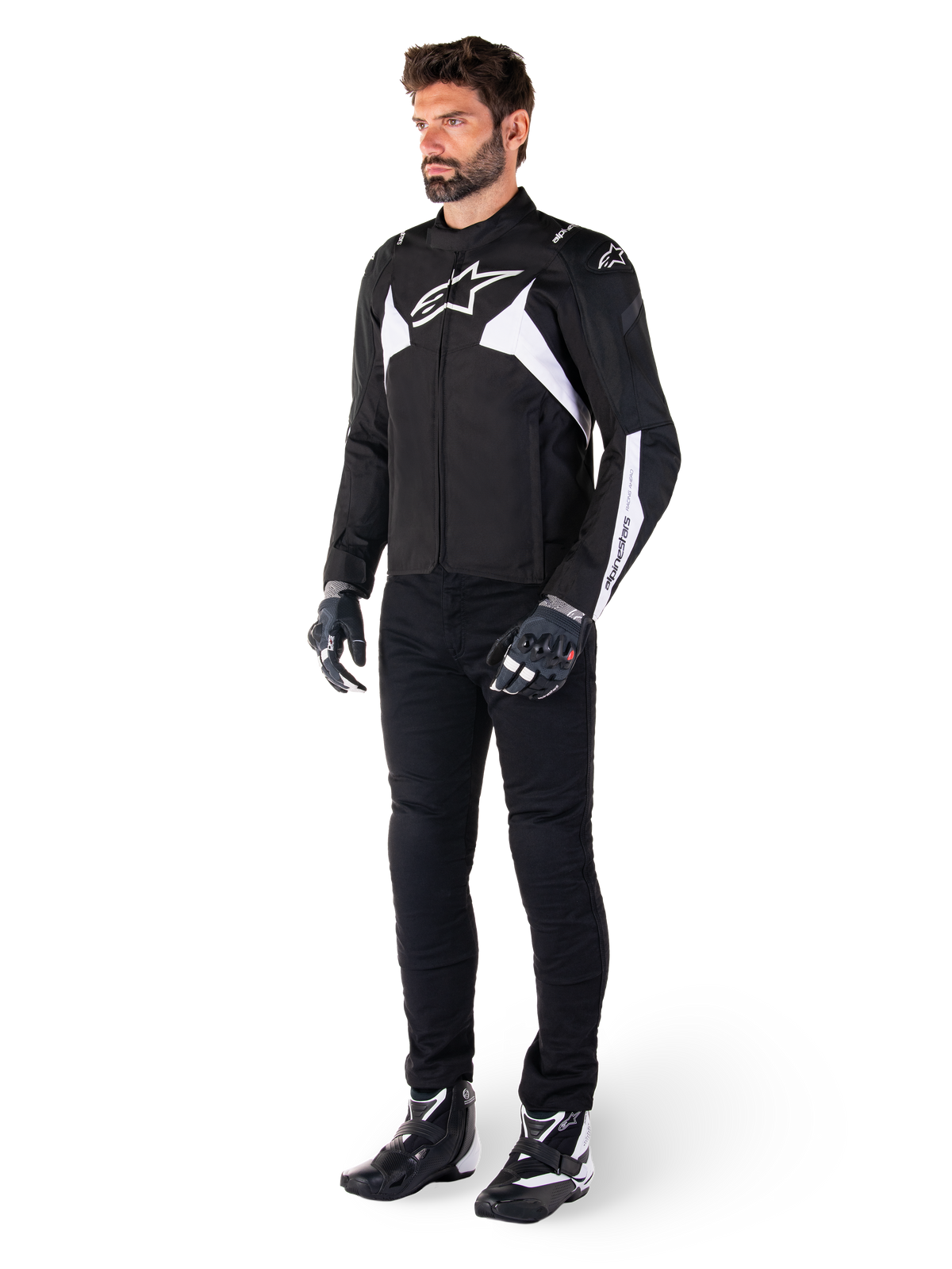 Modelo masculino a usar o Casaco Alpinestars T-Jaws V4 Waterproof em preto e branco, casaco têxtil desportivo com logótipo de estrela branca e detalhes nas mangas, combinado com calças, luvas e botas de motociclismo pretas para um look completo de condução em estrada.