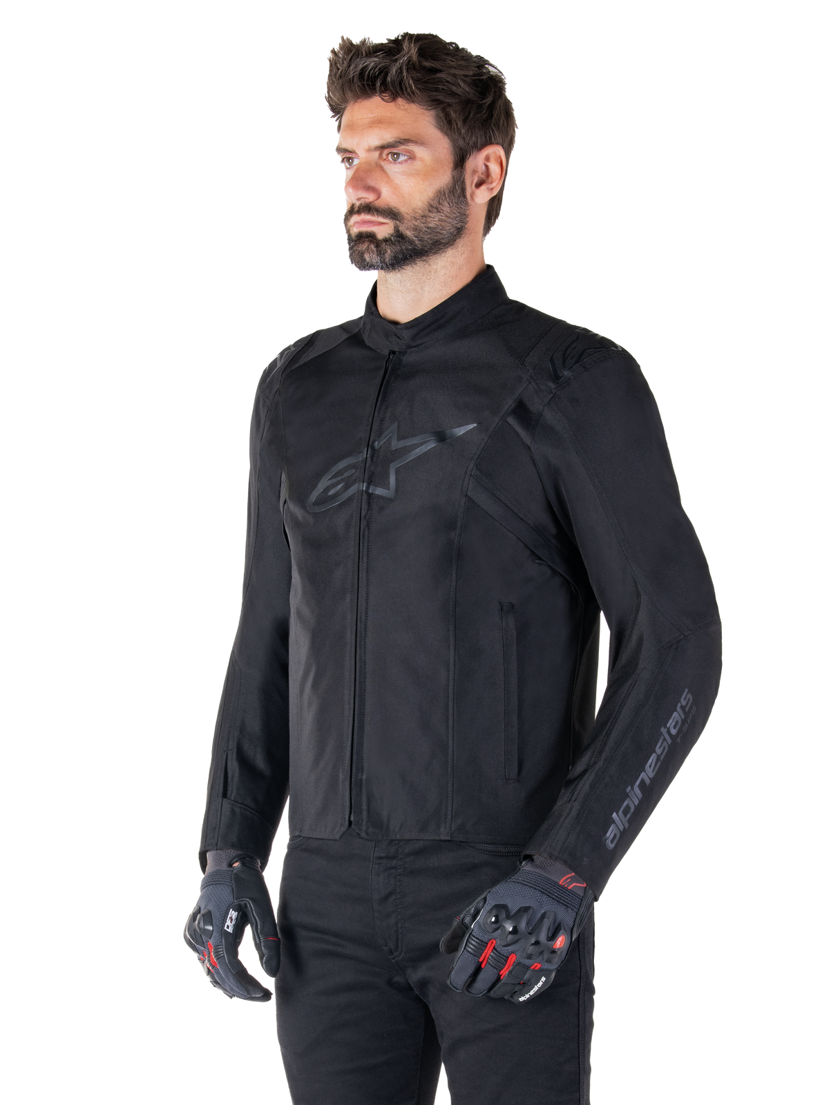Modelo a usar o casaco Alpinestars T-SPS V2 Waterproof, preto, combinado com calças pretas e luvas de motociclismo cinzentas com detalhes vermelhos, com um logótipo preto subtil no peito e marca na manga, casaco têxtil de corte desportivo para condução em estrada