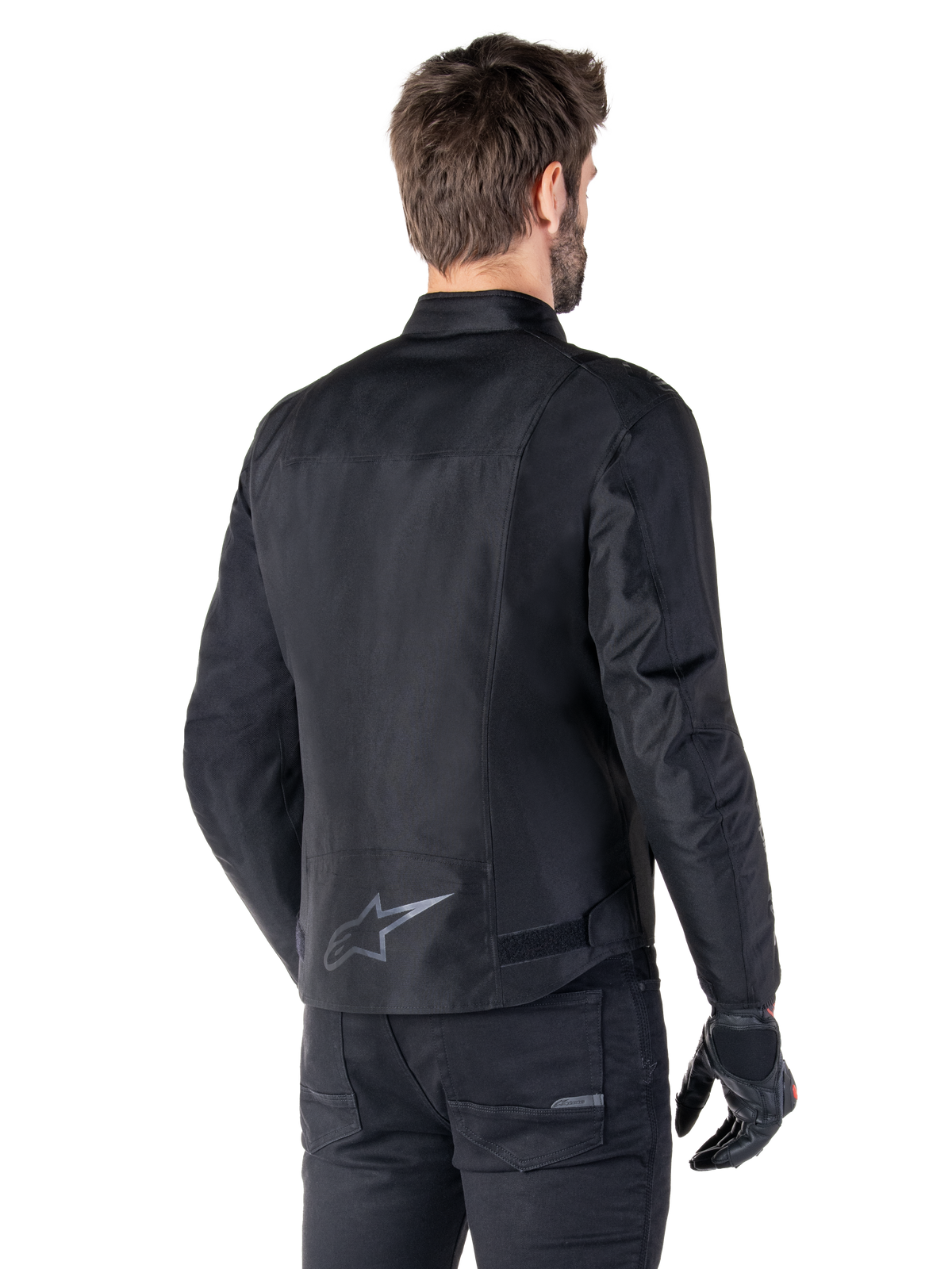 Casaco Alpinestars T-SPS V2 Waterproof, preto, vista traseira num modelo masculino, com logótipo de estrela refletor na parte inferior das costas, perfil traseiro prolongado e fitas de ajuste na cintura, combinado com calças e luvas de condução pretas.
