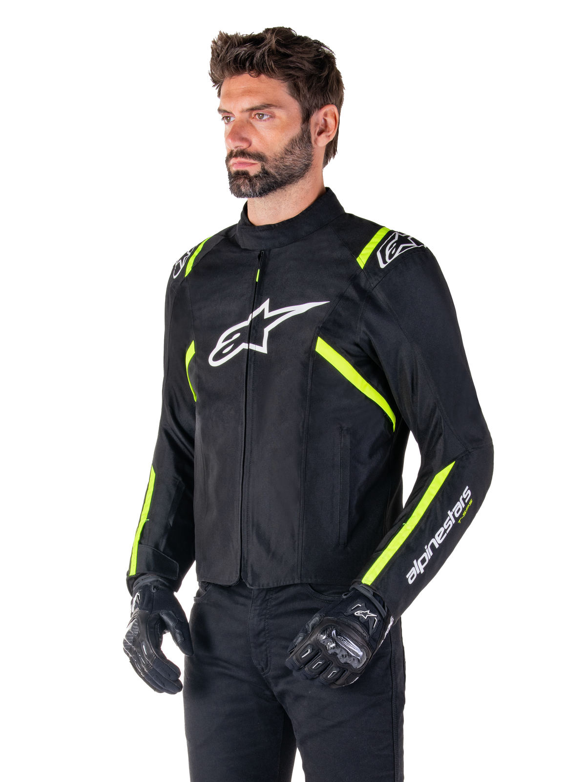 Modelo masculino a usar o casaco Alpinestars T-SPS V2 Waterproof, preto com detalhes em branco e amarelo fluo, com o logótipo Astars branco no peito e riscas amarelo fluorescente nos ombros e mangas, combinado com calças pretas e luvas de motociclismo, casaco de motociclismo têxtil com corte desportivo.