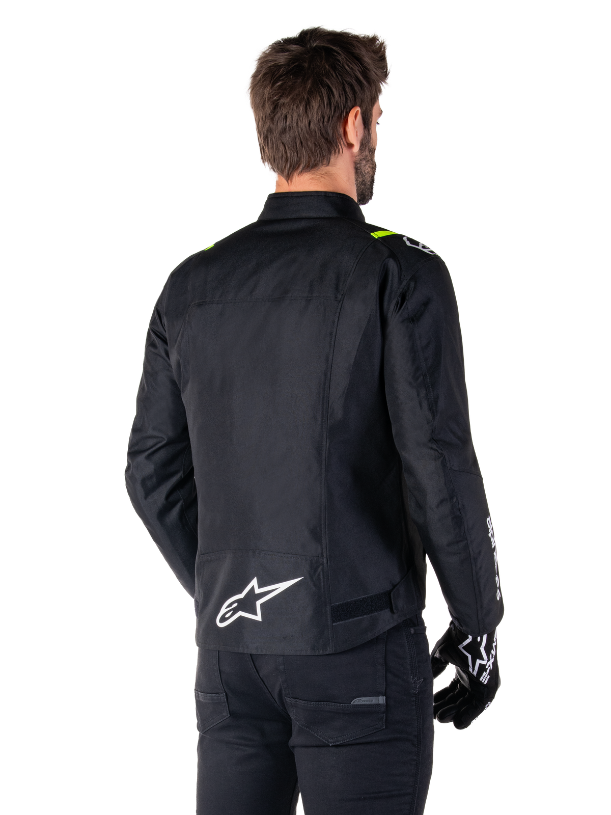 Alpinestars T-SPS V2 Waterproof Jacket, Preto Branco Amarelo Fluo, vista traseira de modelo masculino a usar o casaco de motociclismo têxtil, com logótipo branco na parte inferior das costas e detalhes em amarelo fluo nos ombros, combinado com calças e luvas pretas.