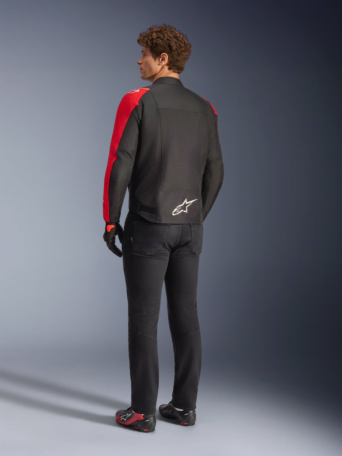 Casaco Impermeável Alpinestars T-SPS V2 em preto e vermelho fluo, vista traseira no modelo, usado com calças de motociclismo pretas e sapatos de condução, com logótipo Alpinestars branco na parte inferior das costas, design de casaco têxtil com corte desportivo