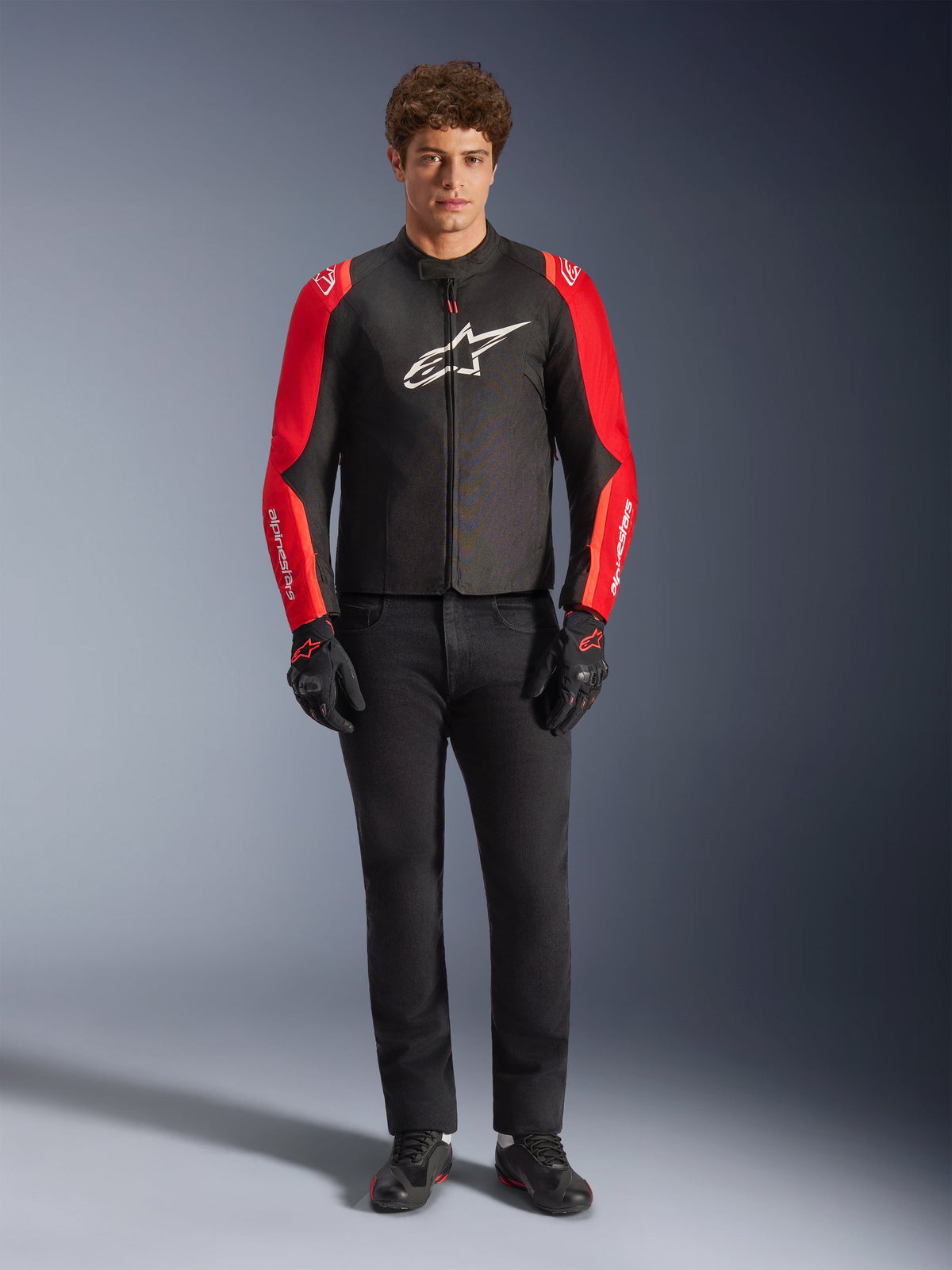 Modelo masculino a usar o Casaco Impermeável Alpinestars T-SPS V2, preto com mangas vermelhas e vermelho fluo, logótipo branco no peito, combinado com calças pretas, luvas e sapatos de condução pretos, design desportivo de equipamento de estrada para motociclismo