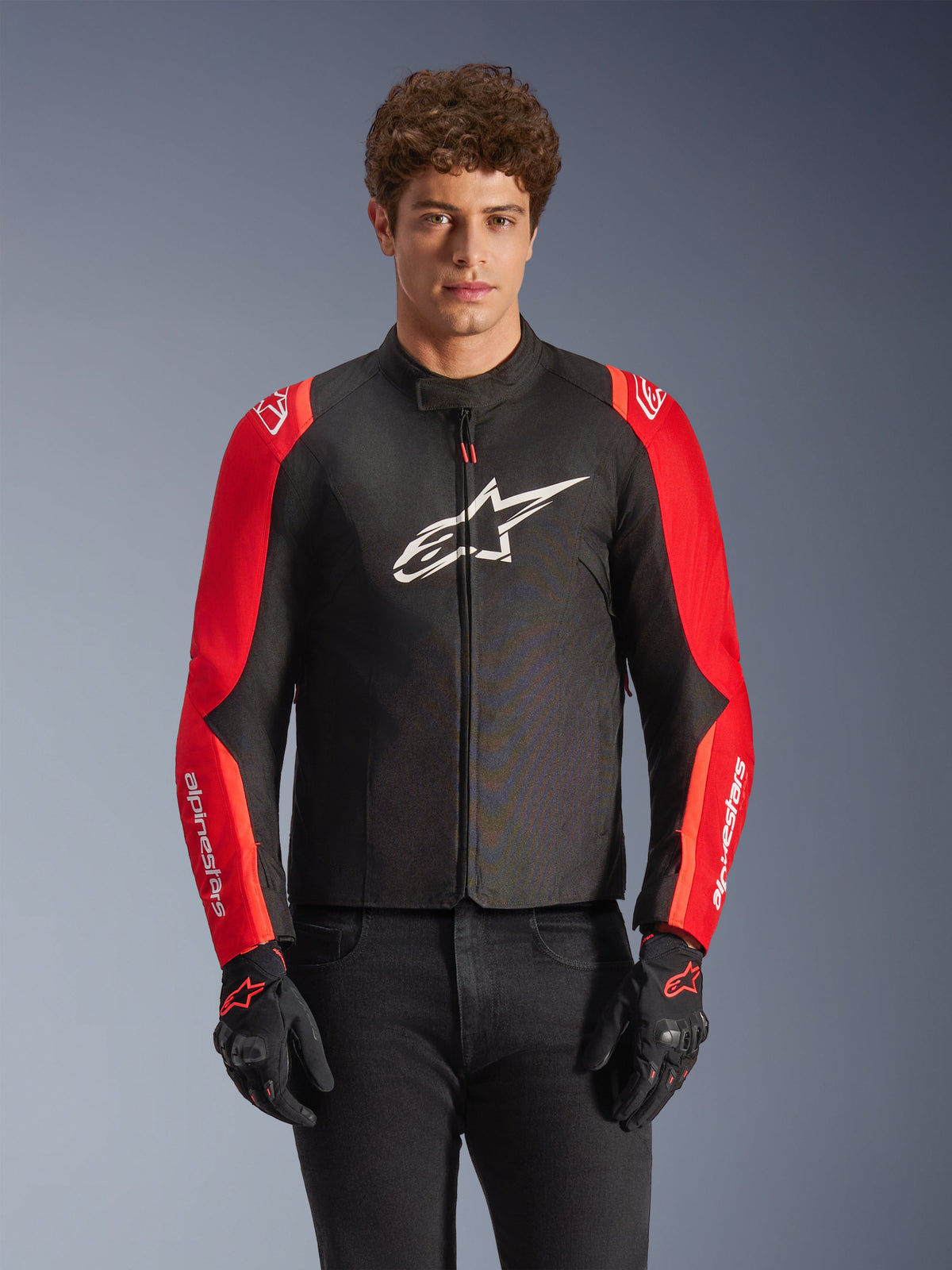 Alpinestars T-SPS V2 Waterproof Jacket, casaco de motociclismo em têxtil, preto e vermelho, usado por um modelo masculino com luvas pretas a condizer, com um logótipo branco grande no peito e marca branca nas mangas vermelhas, silhueta desportiva para condução em estrada em todas as condições climatéricas