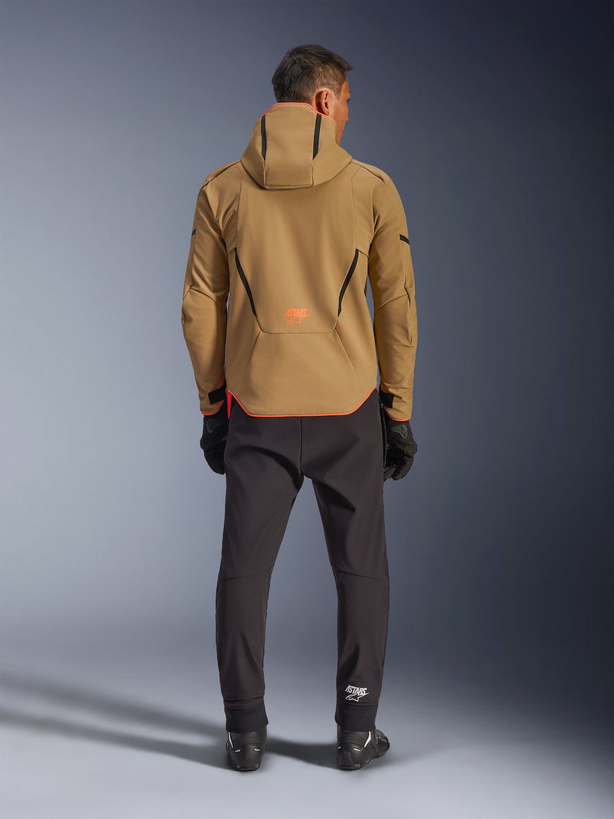 Alpinestars Aeroshell Waterproof Jacket in Dark Khaki Black Orange Fluo, back view of a man wearing the hooded softshell jacket, featuring black piping and orange logo, paired with black technical pants and gloves.