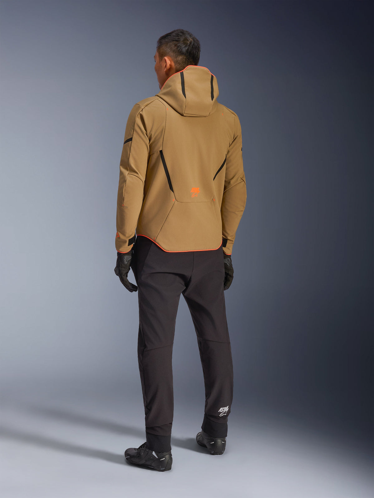 Alpinestars Aeroshell Waterproof Jacket, Dark Khaki Black Orange Fluo, back view of a rider wearing the hooded softshell jacket, paired with black pants and gloves, featuring black geometric accents and an orange logo on the lower back.
