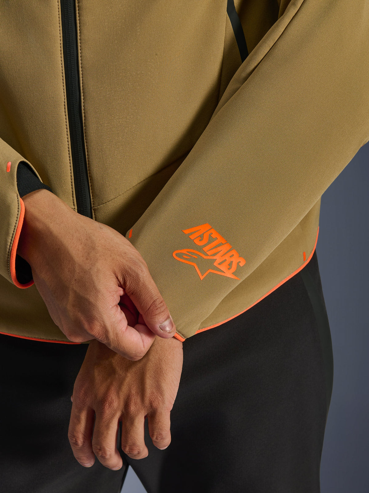 Alpinestars Aeroshell Waterproof Jacket, Dark Khaki with Orange Fluo accents, close-up of the sleeve cuff and lower hem featuring the bright orange Astars logo, neon orange piping, and waterproof front zipper detail, sport softshell construction.