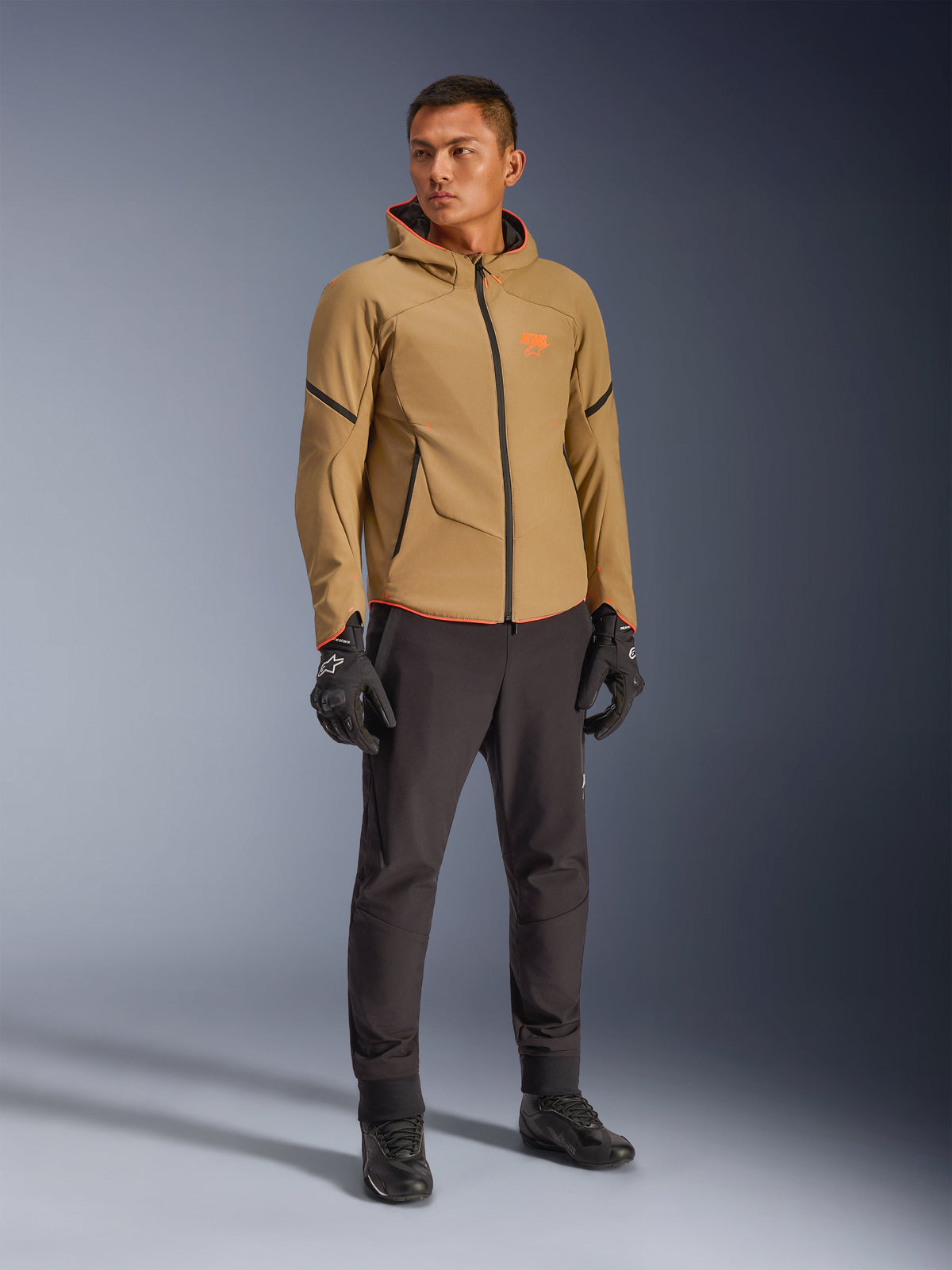 Alpinestars Aeroshell Waterproof Jacket in Dark Khaki, Black, and Orange Fluo, worn by a model with matching gear, featuring a hooded softshell design, waterproof zippers, and bright orange accents and branding.