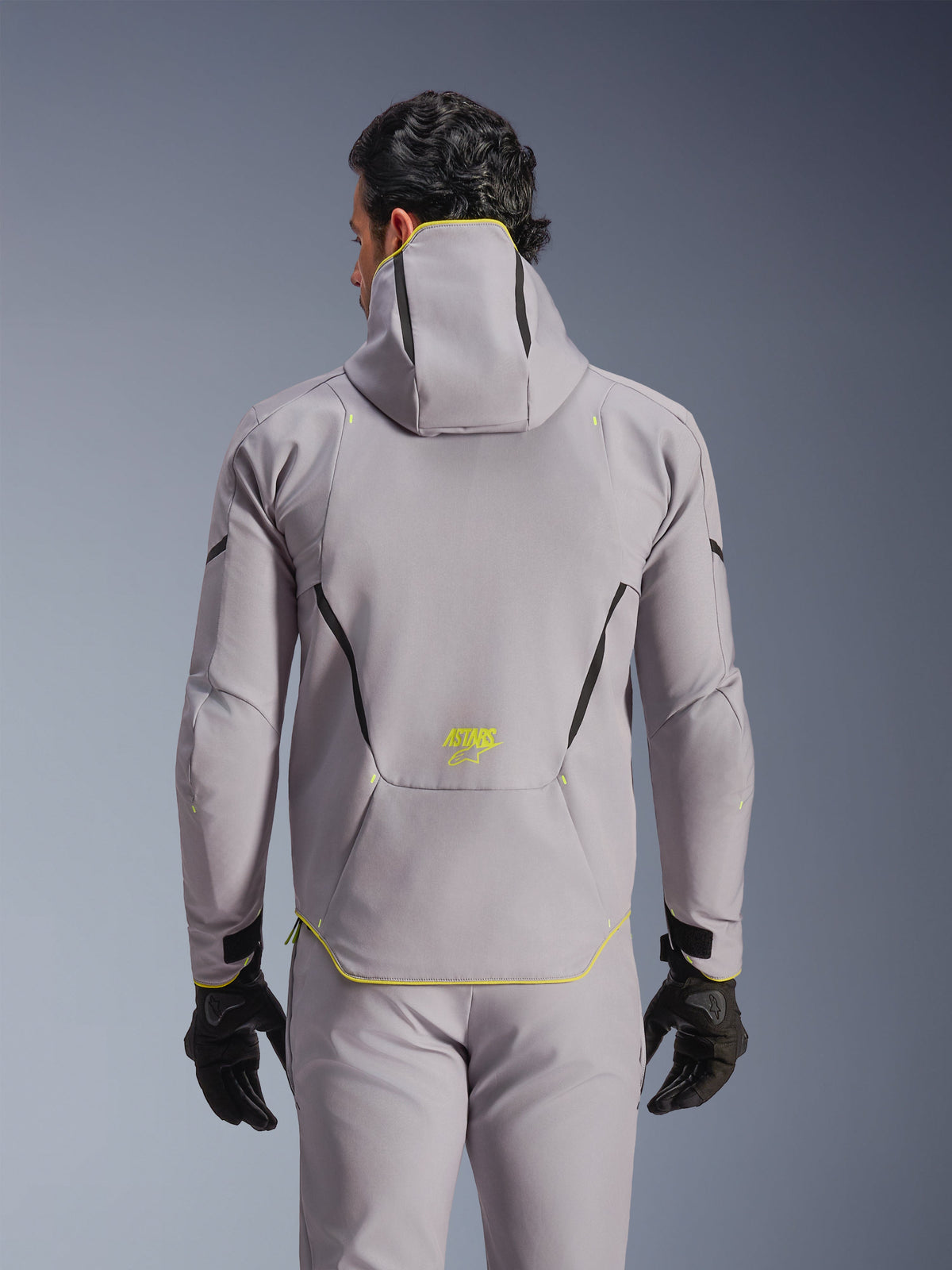 Casaco Alpinestars Aeroshell Waterproof Jacket, cinzento médio com pormenores em amarelo néon, vista traseira mostrando o capuz integrado e linhas de contraste pretas, logótipo ASTARS amarelo na parte inferior das costas, usado com calças cinzentas a condizer e luvas pretas, casaco de motociclismo softshell desportivo
