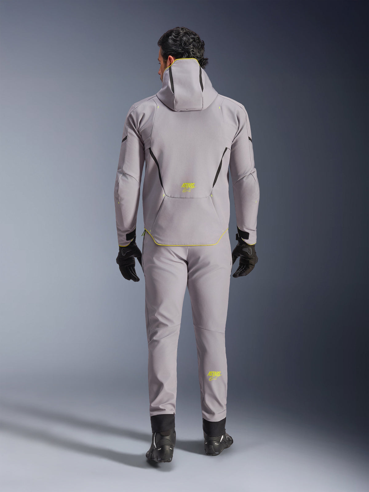 Casaco Alpinestars Aeroshell WP em cinzento e amarelo fluo, vista traseira completa, usado com calças cinzentas a condizer, luvas pretas e sapatos de condução pretos. Apresenta um design softshell com capuz, logótipo 'ASTARS' e detalhes em amarelo fluo, concebido para performance moderna de motociclismo em estrada.