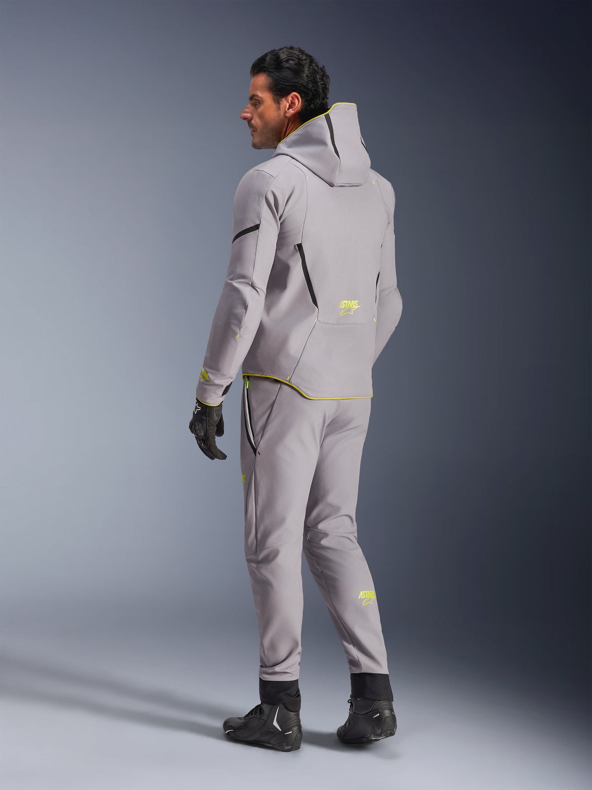 Casaco Impermeável Alpinestars Aeroshell, Cinzento Médio e Amarelo Fluo, usado com calças cinzentas a condizer, com design de hoodie com detalhes e logótipos amarelos, vista de costas, combinado com luvas e botas de motociclismo pretas, vestuário de motociclismo moderno