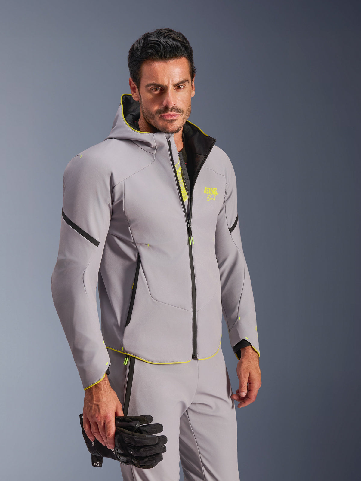 Casaco Alpinestars Aeroshell Waterproof em cinzento médio e amarelo fluo, usado por um homem com calças cinzentas a condizer e luvas de motociclismo pretas, apresentando um design softshell com capuz, pormenores em amarelo néon e logótipo no peito, equipamento de motociclismo desportivo moderno.