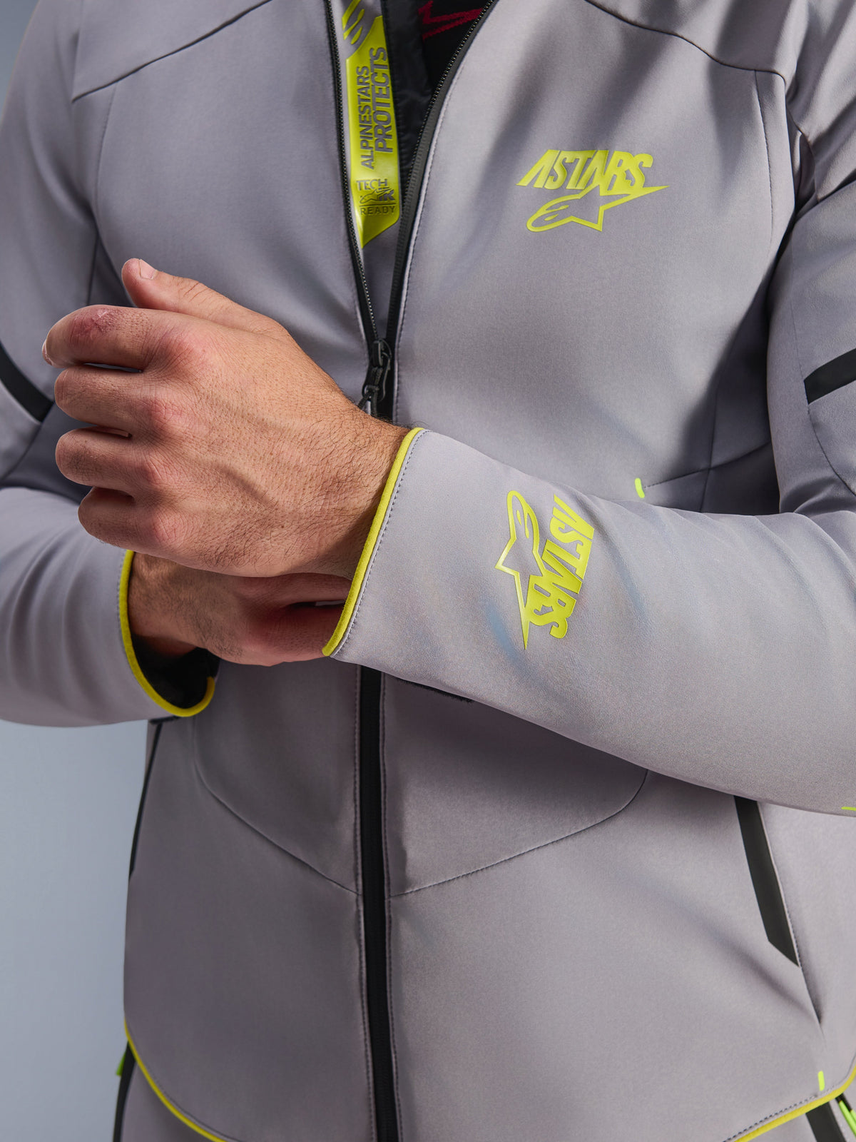 Casaco Impermeável Alpinestars Aeroshell, Cinzento Médio com detalhes em Amarelo Fluo, grande plano do peito e do punho da manga com logótipos Astars em amarelo fluorescente, detalhe da aba interna com marcação Tech-Air Ready, casaco de motociclismo softshell desportivo moderno