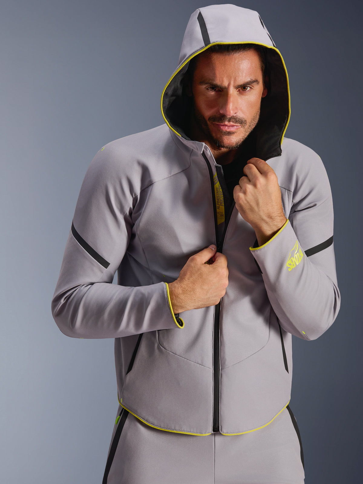 Alpinestars Casaco Aeroshell WP, cinzento médio e amarelo néon, casaco softshell desportivo com capuz, modelo masculino com o capuz levantado, fechos de correr pretos impermeáveis e detalhes em amarelo fluorescente nos punhos e na bainha, design moderno inspirado no desporto motorizado
