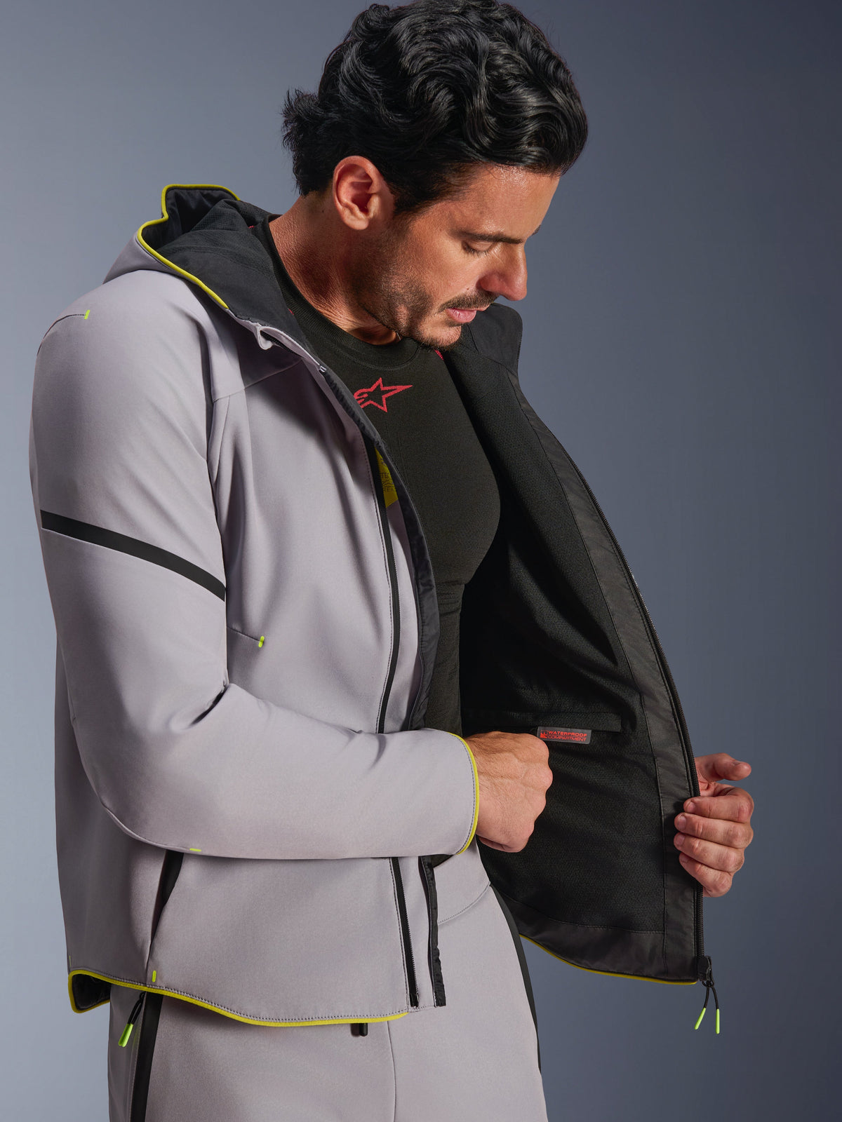 Casaco Alpinestars Aeroshell WP em Cinzento Médio e Amarelo Fluo, vestido por um modelo masculino a mostrar o compartimento interno impermeável e o forro preto, hoodie softshell desportivo para motociclismo com detalhes em amarelo fluorescente e ajuste atlético moderno.