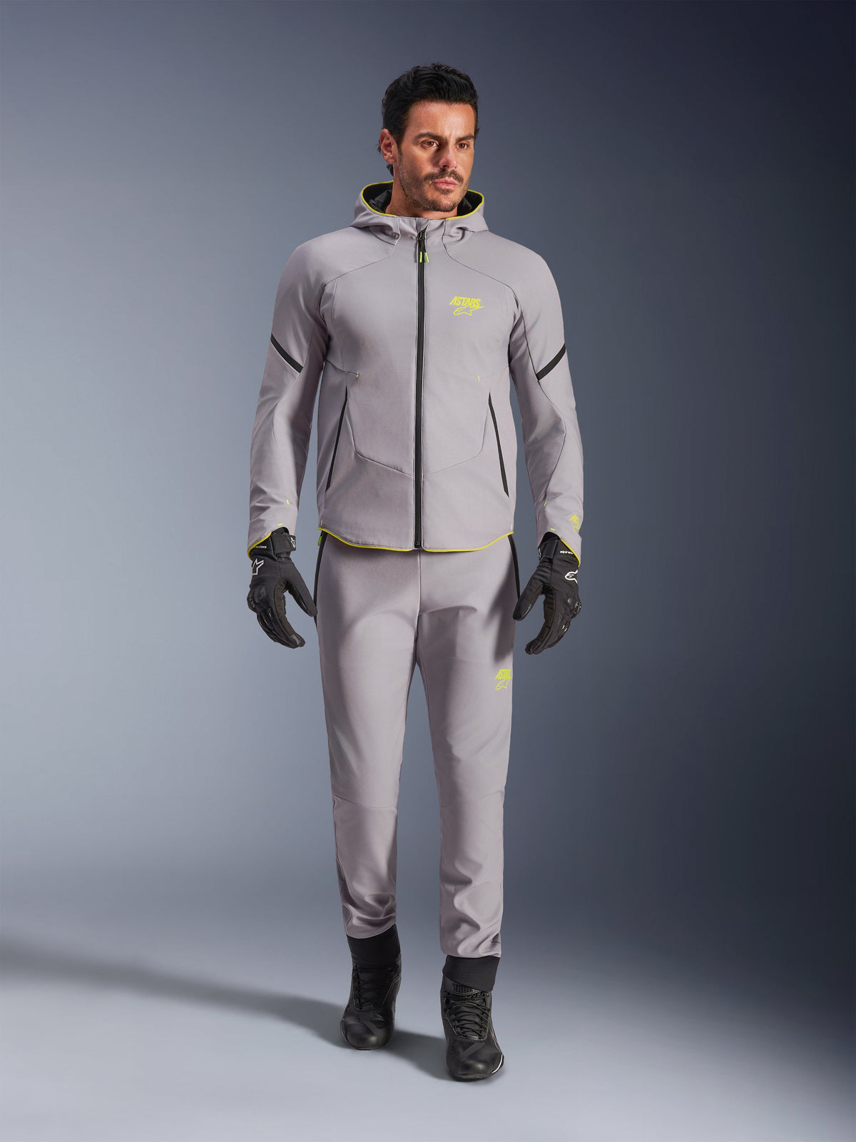 Casaco Alpinestars Aeroshell Waterproof, Cinzento Médio Amarelo Fluo, usado com calças cinzentas a condizer, luvas e botas pretas, com um design de hoodie moderno inspirado no desporto, detalhes em amarelo vibrante e um corte atlético.