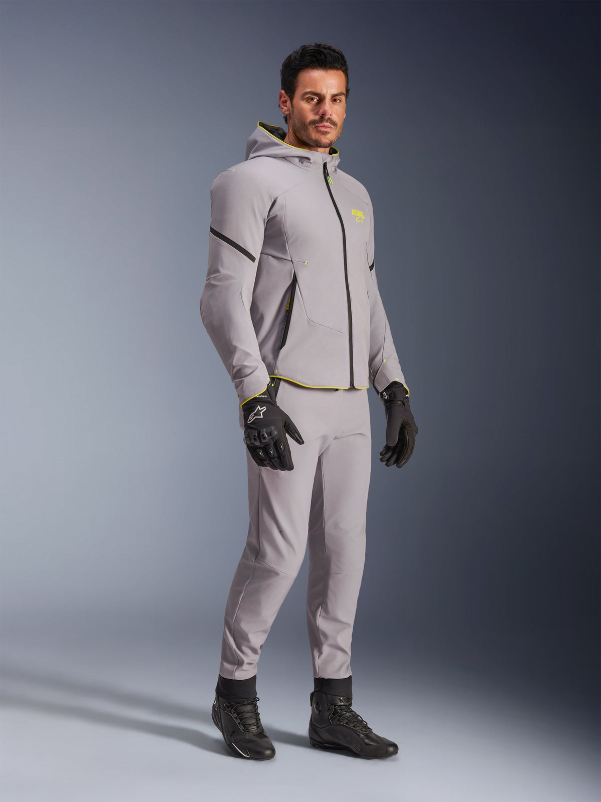 Casaco Alpinestars Aeroshell WP, cinzento médio e amarelo fluo, usado por modelo com calças cinzentas a condizer, luvas de motociclismo pretas e botas pretas, equipamento de motociclismo moderno tipo hoodie para condução casual e desportiva.