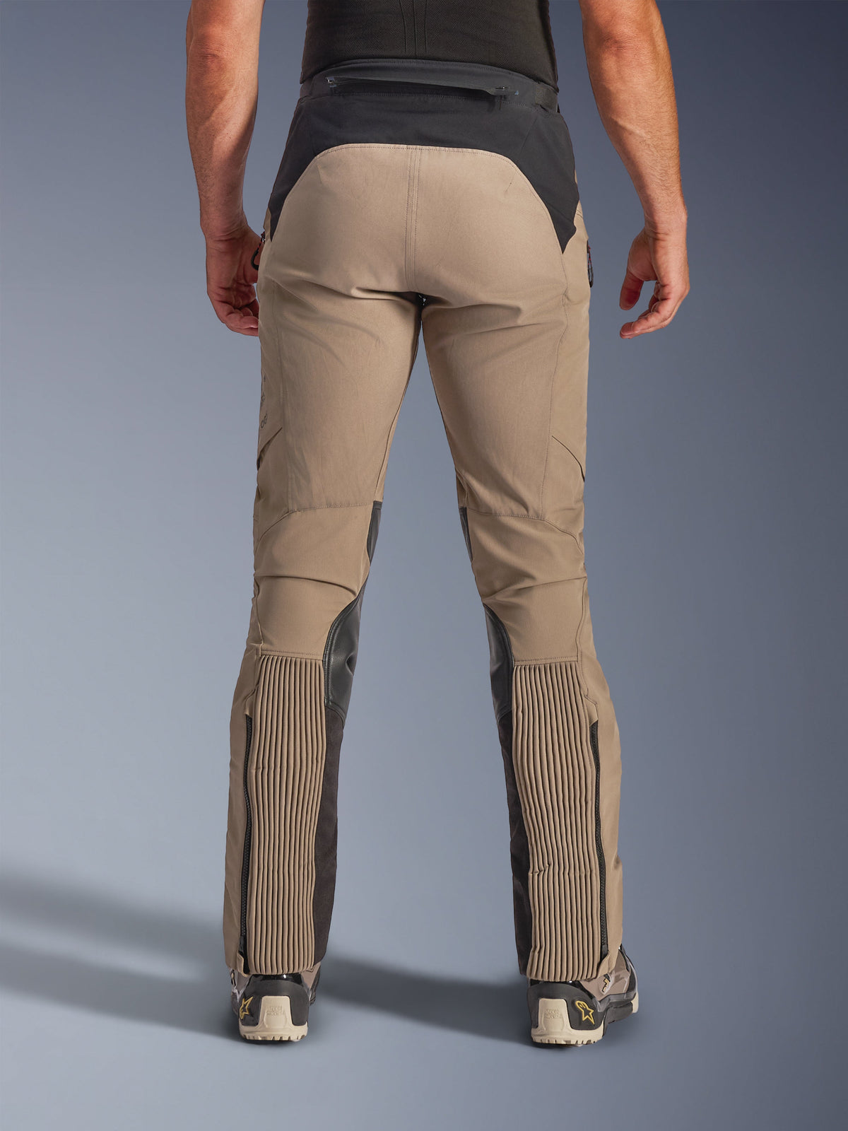 Calças Alpinestars AMT-8 Stretch Drystar® XF, Walnut Black, vista traseira no modelo, base têxtil elástica em bege com escapulário preto na cintura e painéis na barriga da perna, com grandes secções em acordeão elástico na parte inferior das pernas, entradas das pernas com fecho de correr e corte adventure-touring