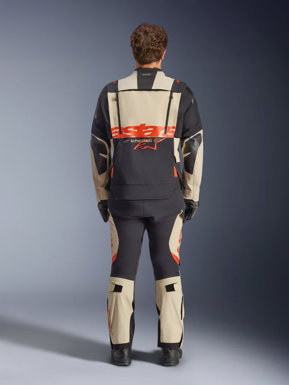 Casaco laminado Alpinestars Halo Pro Drystar® XF, casaco de motociclismo de aventura, Mastic Black com logótipo cor de laranja, vista traseira que mostra o logótipo grande nas costas e fechos de ventilação modulares, usado com calças de aventura a condizer para desempenho em todo-o-terreno