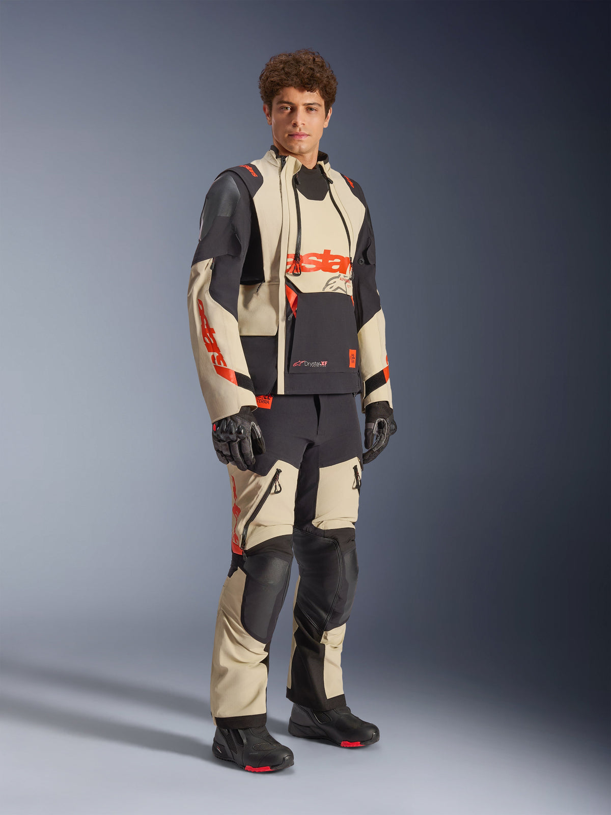 Modelo masculino a usar o Casaco Alpinestars Halo Pro Drystar® XF Laminated em Mastic Black, com painéis bege e pretos e detalhes em vermelho, combinado com calças de aventura a condizer e luvas pretas, concebido para motociclismo de aventura em todo-o-terreno.