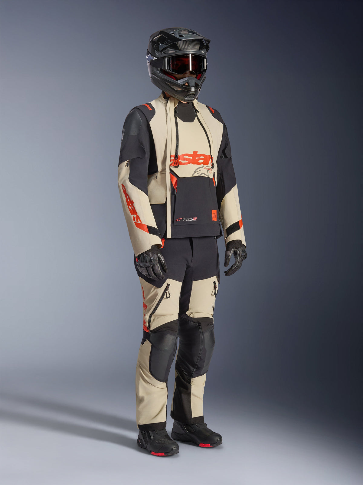 Alpinestars Halo Pro Drystar® XF Laminated Jacket em Mastic Black, casaco de motociclismo de aventura usado com calças a condizer, capacete integral, luvas e botas, apresentando painéis bege e pretos com logótipos vermelhos, design técnico multiestação