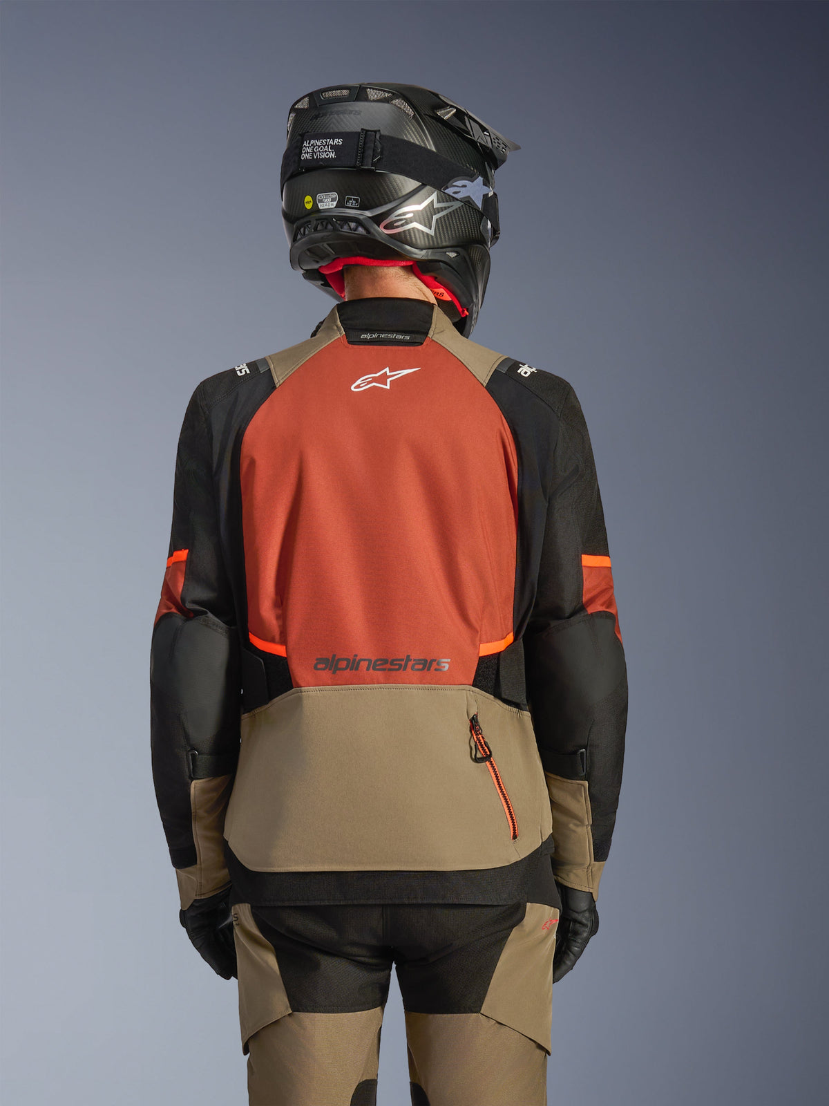 Vista traseira do Casaco Alpinestars Andes V4 Drystar® em Walnut Rust Black, casaco de aventura e touring com um grande painel traseiro cor de ferrugem com logótipo branco, secção inferior em nogueira com bolso de fecho vertical, mangas pretas com detalhes em laranja, combinado com calças a condizer e um capacete preto para condução em qualquer clima