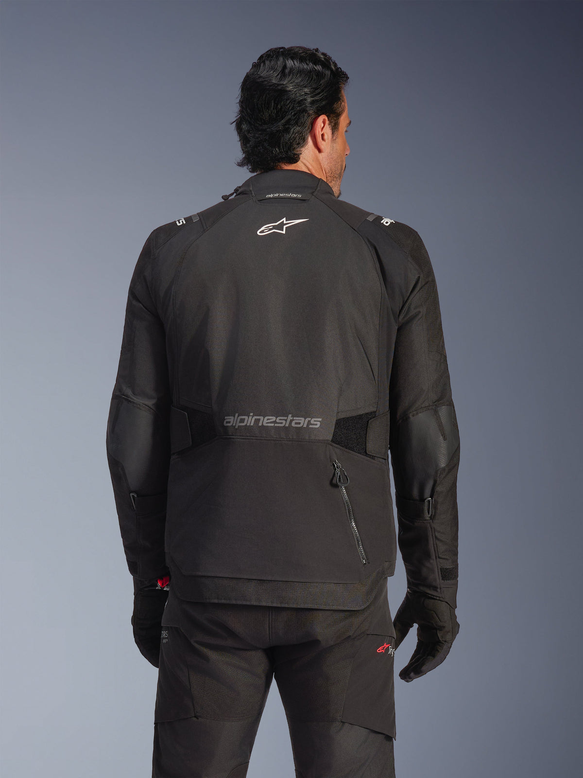 Casaco Alpinestars Andes V4 Drystar®, preto, vista traseira num modelo, casaco têxtil de aventura com logótipos refletores e um bolso traseiro com fecho de correr, combinado com calças e luvas pretas a condizer