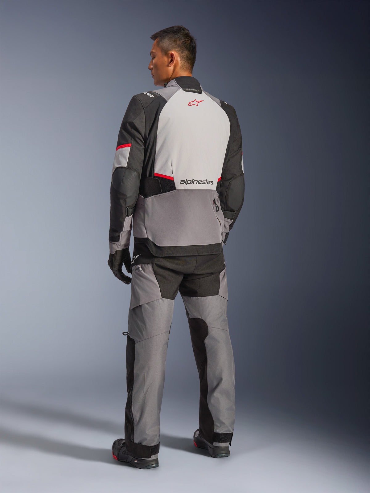 Casaco Alpinestars Andes V4 Drystar®, cinzento escuro e cinzento gelo com detalhes em preto e vermelho vivo, vista traseira mostrando painel cinzento claro com logótipo de estrela vermelho, combinado com calças de aventura a condizer e luvas pretas, equipamento técnico de motociclismo para todas as condições meteorológicas.