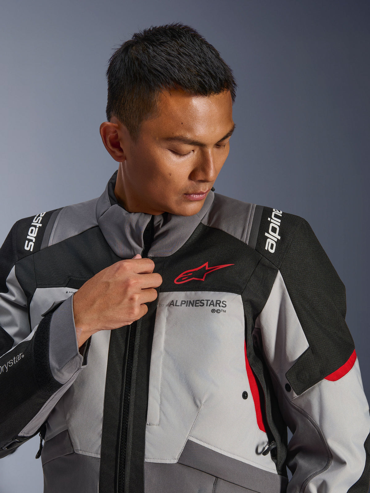 Modelo a usar o Casaco Alpinestars Andes V4 Drystar®, cinzento escuro e cinzento gelo com detalhes em preto e vermelho vivo, a ajustar o fecho frontal, com o logótipo Astars vermelho e marca Drystar®, estilo adventure touring
