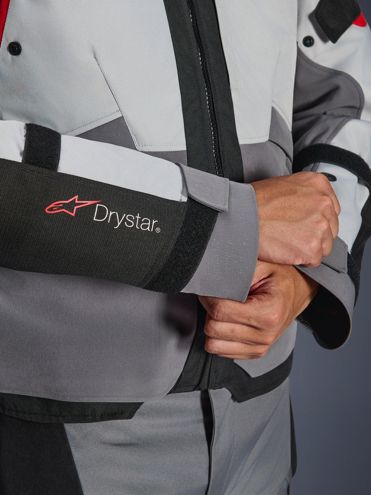 Detalhe em grande plano da manga do casaco Alpinestars Andes V4 Drystar®, têxtil cinzento e preto, com o logótipo Drystar vermelho e branco no antebraço, fecho de velcro ajustável no pulso, equipamento de motociclismo impermeável de alta performance