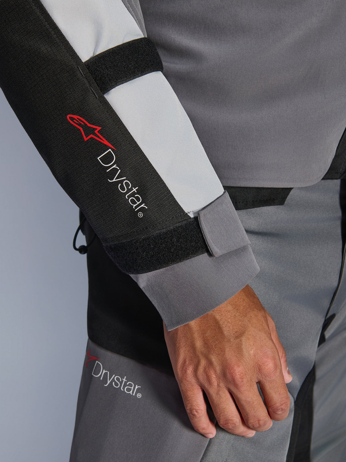 Pormenor da manga do casaco Alpinestars Andes V4 Drystar®, cinzento escuro e cinzento gelo com detalhes em preto, destacando o logótipo Drystar® e as correias de ajuste no antebraço, parte de um equipamento têxtil de motociclismo de aventura impermeável