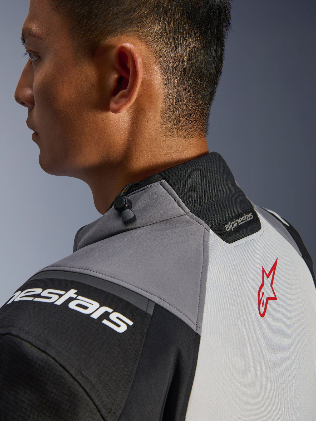 Casaco Alpinestars Andes V4 Drystar®, grande plano da gola e da parte superior das costas, Cinzento Escuro e Cinzento Gelo com detalhes pretos, logótipo de estrela vermelha, inclui ajustador de gola por cordão e marca Alpinestars, construção têxtil durável