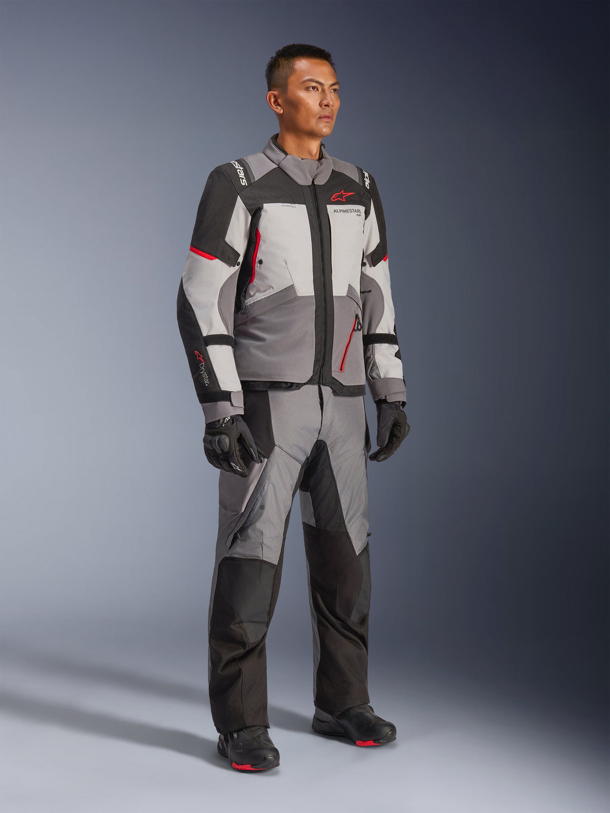 Modelo masculino a usar o Casaco Alpinestars Andes V4 Drystar® e calças de aventura a combinar, em Cinzento Escuro, Cinzento Gelo, Preto e Vermelho Brilhante, com luvas e botas de motociclismo pretas, equipamento técnico para todas as condições meteorológicas com design moderno de vários painéis.