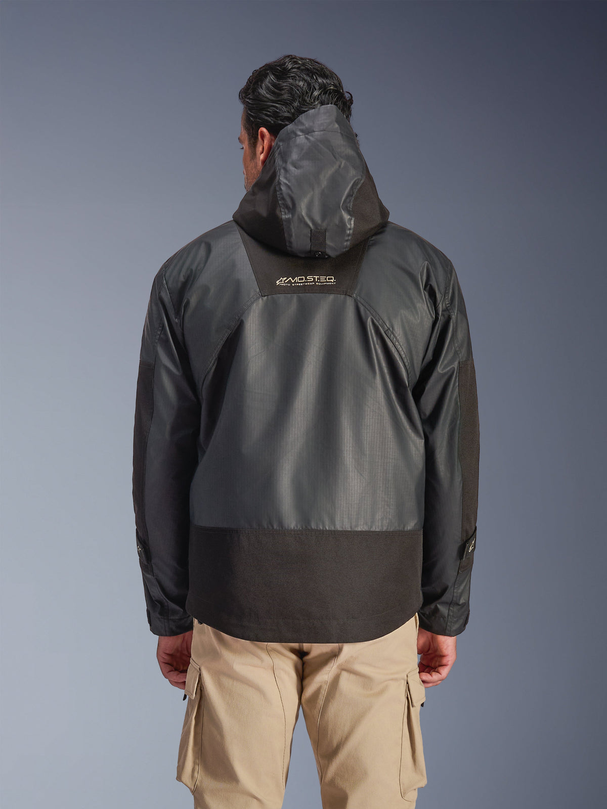 Vista traseira de um homem a usar o Casaco Impermeável Alpinestars Mospher em preto, com capuz e construção durável em tecido ripstop, combinado com calças cargo beges, realçando o logótipo MO.ST.EQ. na parte superior das costas, design de equipamento de motociclismo urbano.