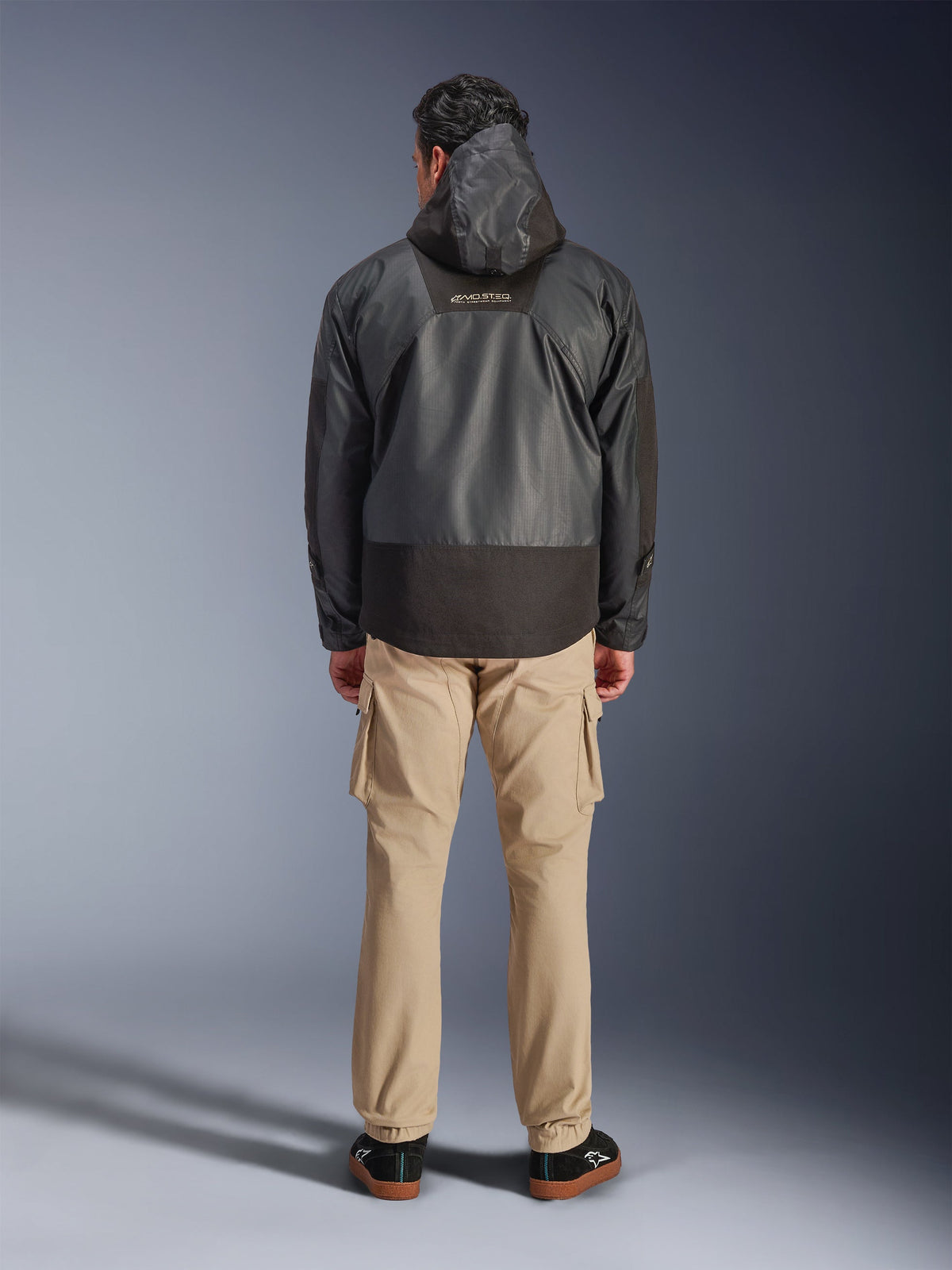 Alpinestars Mospher Waterproof Jacket, parka de motociclismo preta vista de costas, com o logótipo MO.ST.EQ na parte superior das costas e construção em tecido ripstop, usada com calças cargo beges e sapatilhas pretas, estilo urbano de commuting