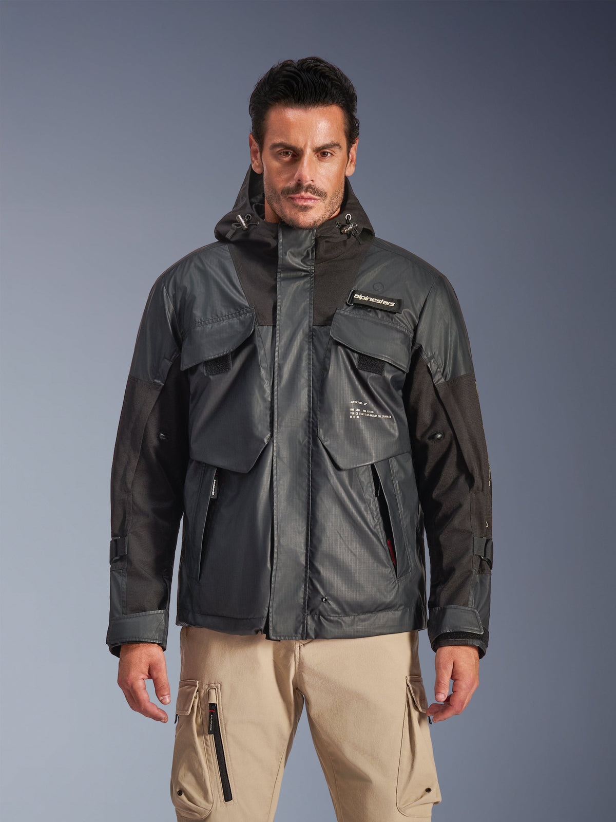 Alpinestars Mospher Waterproof Jacket, parka de motociclismo urbana para homem, tecido ripstop preto e cinzento escuro, design com capuz e múltiplos bolsos utilitários grandes, usada por um modelo com calças cargo caqui para um visual tático.